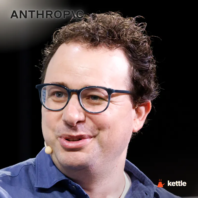 A curly-haired man with glasses in a blue shirt, with the ANTHROPIC logo overlaid, speaking with a microphone visible
