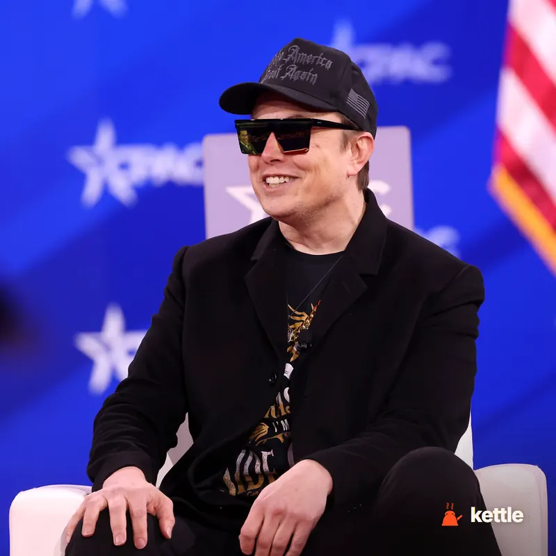 Elon Musk wearing a MAGA cap and sunglasses, smiling, seated at a CPAC event with American flag backdrop