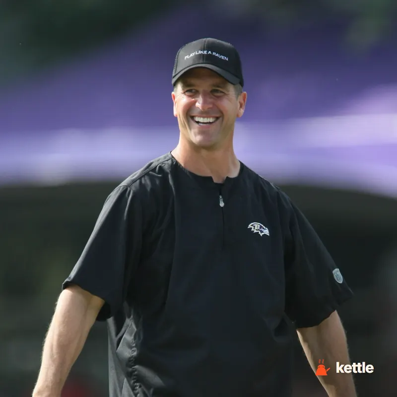 A smiling man in a black Baltimore Ravens coaching cap and jacket, outdoors at a practice field