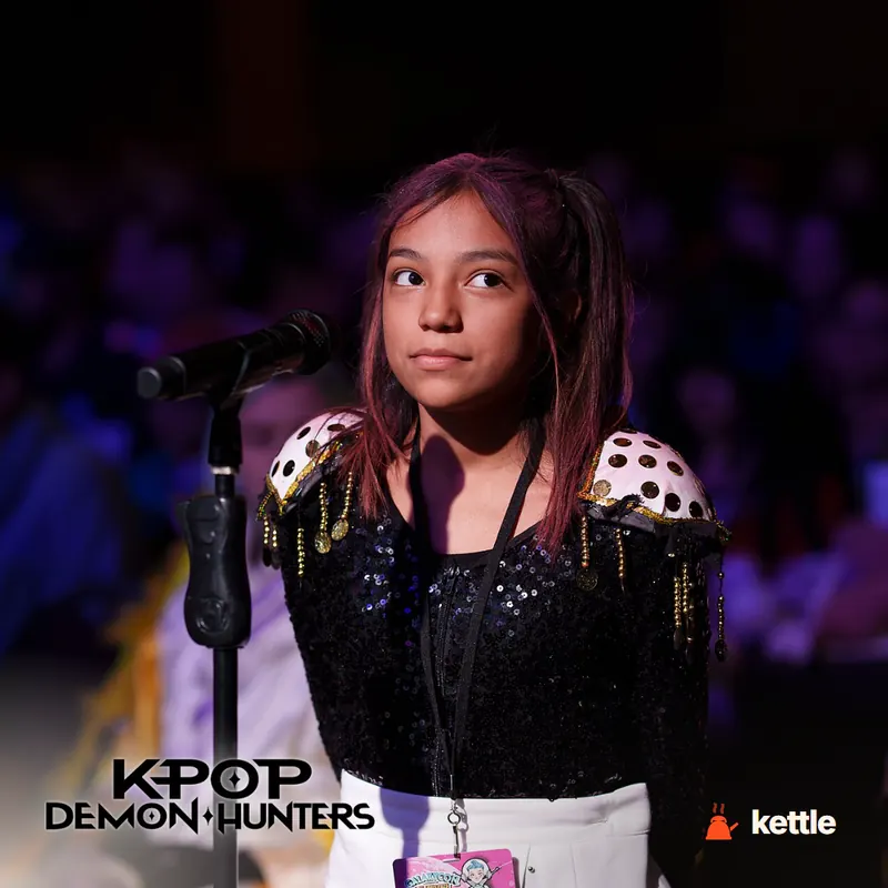 A young girl in costume at a microphone/stage event with the 'KPOP DEMON HUNTERS' wordmark logo overlaid in bottom left corner