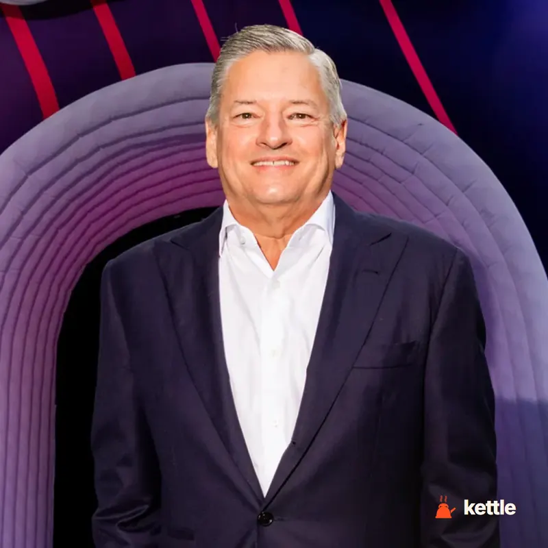 A smiling man in a dark suit at an event with a purple arch backdrop, presumably Ted Sarandos