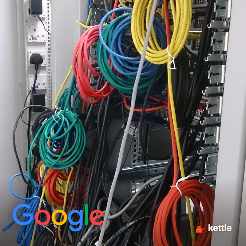A server rack with tangled colorful network cables, with the Google wordmark logo overlaid in the bottom-left corner
