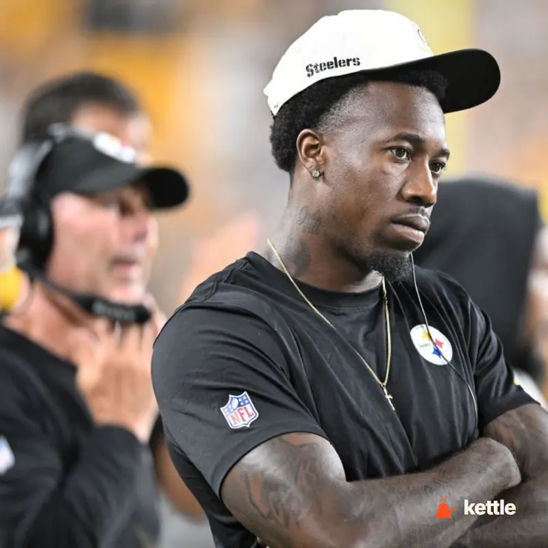 A player wearing a Steelers cap and black Steelers shirt with NFL patch, standing on the sideline with arms crossed, next to a coach with headset